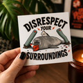 A hand holds a Cool Shiz Neil the seal waterproof, scratch resistant, vinyl, 'DISRESPECT YOUR SURROUNDINGS' sticker. The background shows a warm indoor setting with a lit candle, a wooden surface, and blurred green foliage.