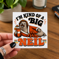 A square white sticker held by a hand, featuring a Cool Shiz Neil The Seal 'I'M KIND OF A BIG NEIL' waterproof, scratch resistant, vinyl sticker. The sticker is held over a wooden table background with a blurred plant and candle.