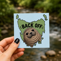 A hand holds a Cool Shiz, Neil the seal, Back Off, Waterproof, Scratch Resistant, Vinyl Sticker, with a blurred outdoor stream and greenery behind it.
