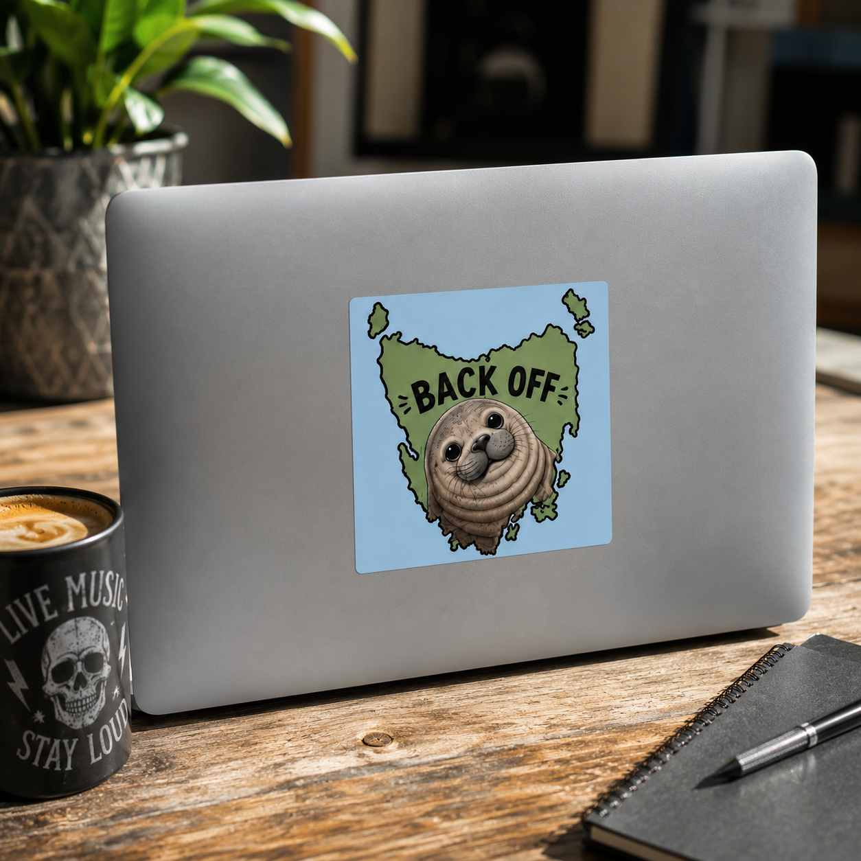 A square sticker featuring a Cool Shiz, Neil the seal, Back Off, Waterproof, Scratch Resistant, Vinyl Sticker on a laptop, on a wooden desk with a mug, notebook, and plant nearby.
