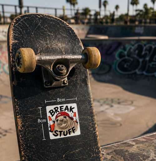 A close-up of a worn black skateboard deck with a Cool Shiz, Neil the seal, Break Stuff, Waterproof, Scratch Resistant, Vinyl Sticker on it. The skateboard is photographed outdoors at a skatepark with ramps and graffiti in the blurred background.