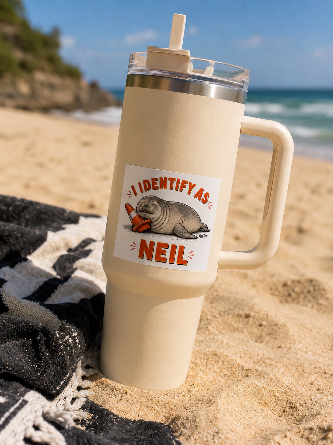 A yellow insulated travel mug on a sandy beach. The mug has a Cool Shiz Neil the seal I Identify As Neil waterproof, scratch resistant, vinyl sticker on it.