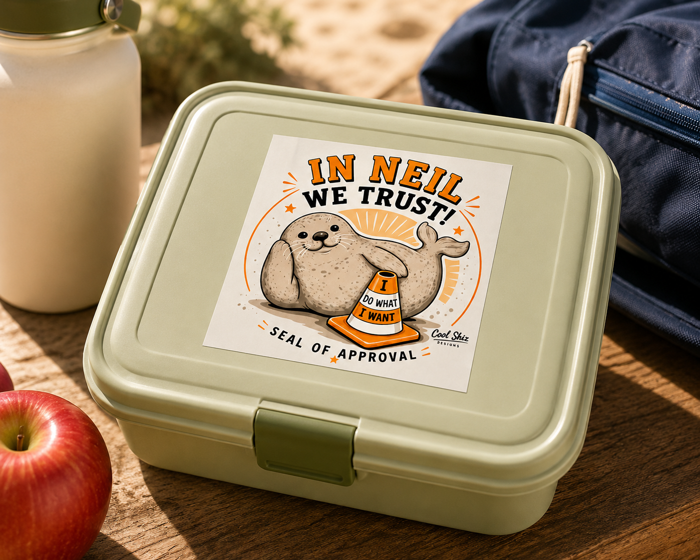 A square bento lunch box featuring a Cool Shiz Neil the seal In Neil We Trust waterproof, scratch resistant, vinyl sticker on the lid. The lunch box is styled on a wooden picnic table with a red apple, a white bottle, and a navy blue backpack in the background.