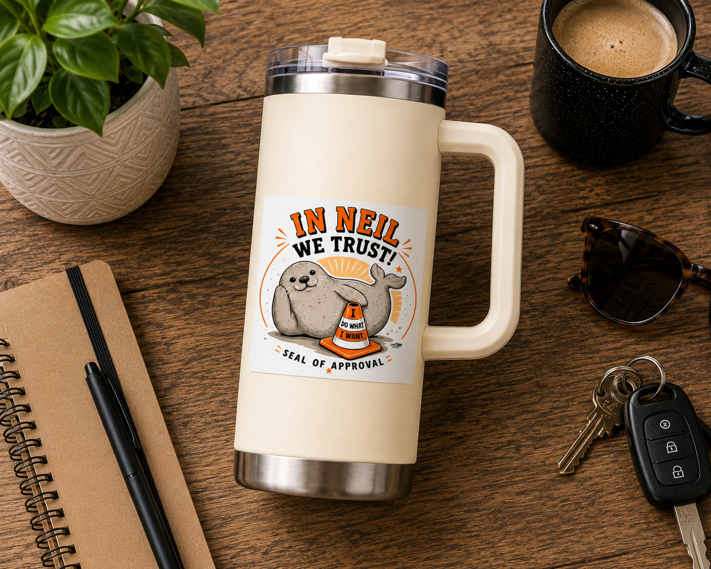 A travel mug featuring a Custom Cool Shiz, Neil the seal, In Neil we trust, 8x8cm, kiss cut, scratch resistant waterproof vinyl sticker. Displayed on a wooden desk surrounded by a notebook, plant, coffee mug, sunglasses and keys.