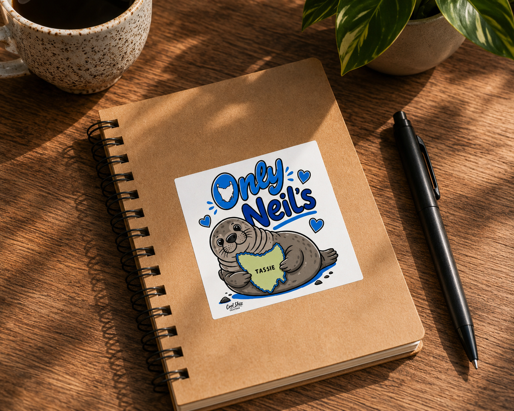 A kraft paper spiral notebook on a wooden desk with a black pen, a speckled mug, and a plant. The notebook cover features a Cool Shiz Neil the seal 'Only Neil's', waterproof, scratch resistant, vinyl sticker.