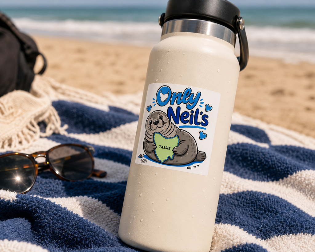 A cream-colored insulated water bottle featuring a Cool Shiz Neil the seal 'Only Neil's', waterproof, scratch resistant, vinyl sticker. The bottle is placed on a blue and white striped beach towel on the sand, with sunglasses and a bag in the background.